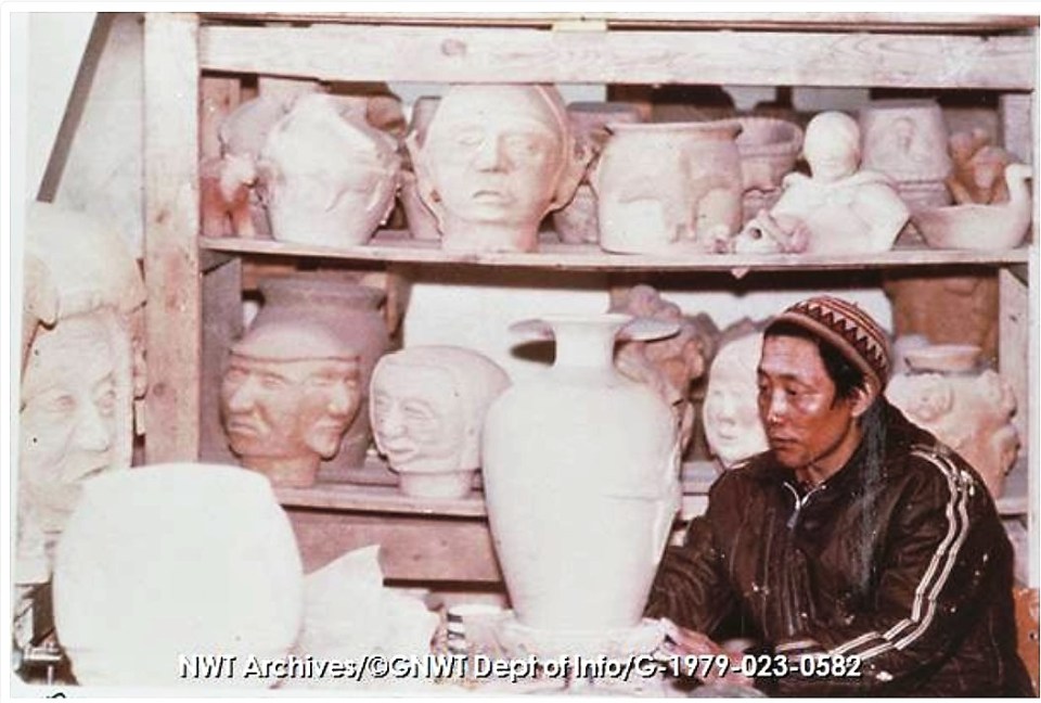Ceramic shop, Rankin Inlet [man sits at table in pottery studio with large urn on pedestal, surrounded by large pots, heads and other figures on shelves ca. 1971]. CREDIT: NWT Archives/Northwest Territories. Department of Information fonds/G-1979-023: 0582. From the holdings of the NWT Archives.